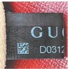 Close view of a Gucci black fabric label showing part of its wordmark and printed code beside red leather
